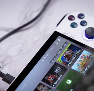 A handheld gaming console with a white body and colorful buttons features a screen displaying various game icons. The device is connected to a power source via a black cable and rests on a marble surface. The interface shows games like Apex and Street Fighter.